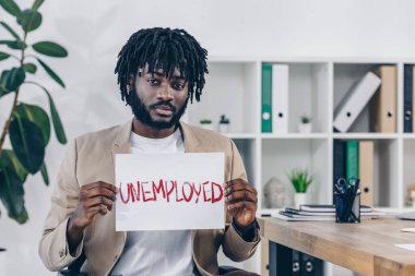 African american employee looking at camera and presenting placard with unemployed lettering in office clipart
