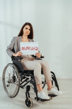 Kameraya bakan engelli bir kadın ve elinde beyaz tekerlekli sandalyede işsiz harfleri olan bir pankart var. 