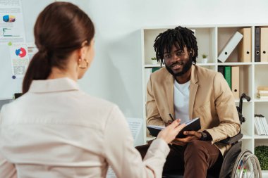 Selective focus of recruiter and african american disabled employee with copybook looking at each other at job interview clipart