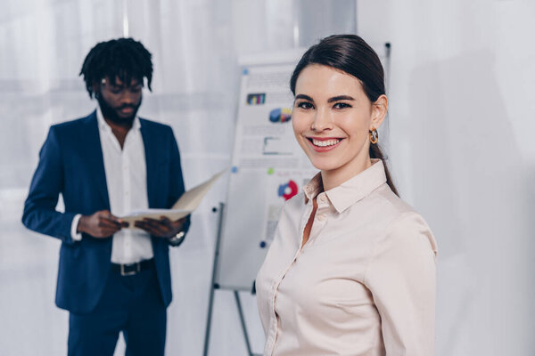 Рекрутер улыбается и смотрит на камеру и африканского американского работодателя возле флип-чарта с папкой в офисе
