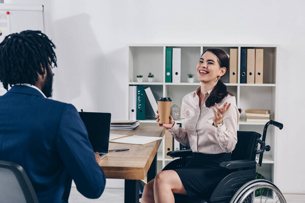 Инвалид на инвалидной коляске с бумажной чашкой кофе смеется рядом с африканским американским рекрутером с ноутбуком
