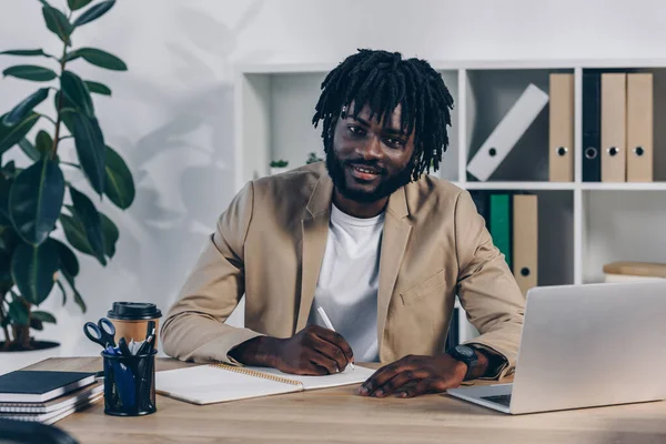 Afrikanischer Rekrutierer Schreibt Notizbuch Neben Laptop Lächelt Und Blickt Die — Stockfoto
