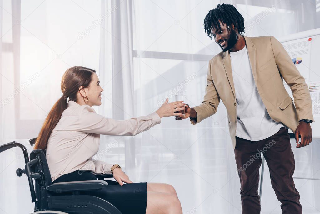 Selective focus of african american employee giving glass of water to disabled recruiter on wheelchair in office