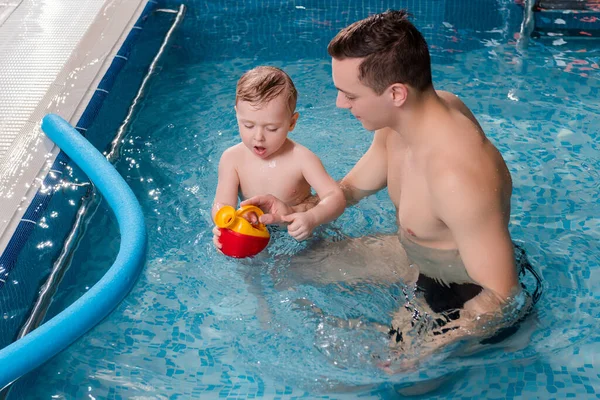swim trainer looking at toddler boy playing in swimming pool