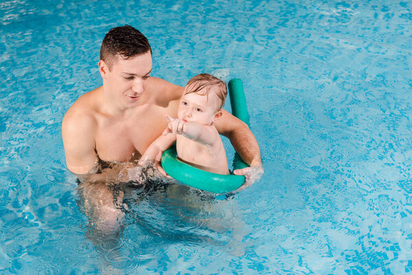 handsome trainer looking at cute toddler boy pointing with finger in swimming pool 