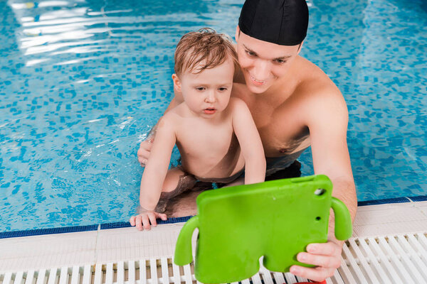 happy swim coach and toddler boy swimming and looking at digital tablet 