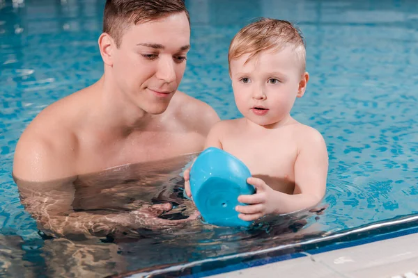 swim coach looking at toy ship in hands of toddler boy in swimming pool