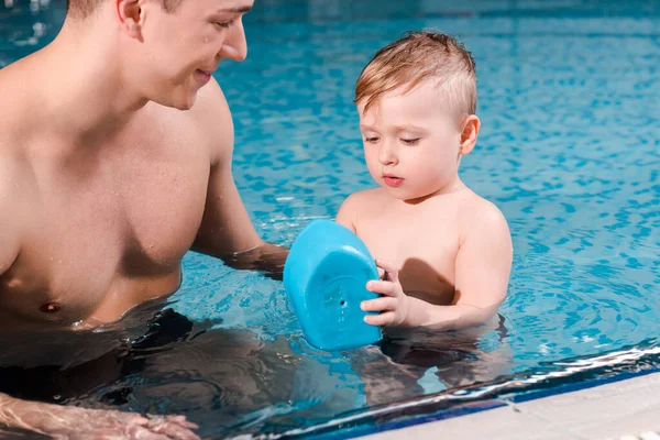 快乐的游泳教练看着蹒跚学步的男孩在游泳池玩具船 — 图库照片