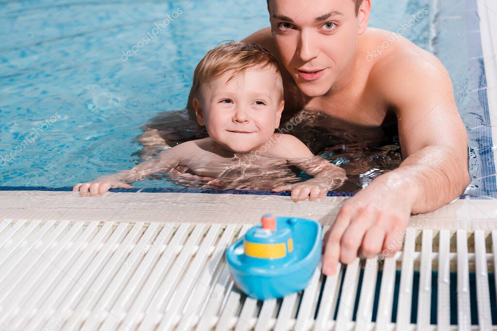 swim coach near happy toddler boy and toy ship in swimming pool 