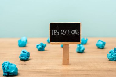 Üzerinde testosteron harfi olan isim levhası, mavi üzerine izole edilmiş ahşap yüzey üzerine buruşmuş kağıdın yanında.