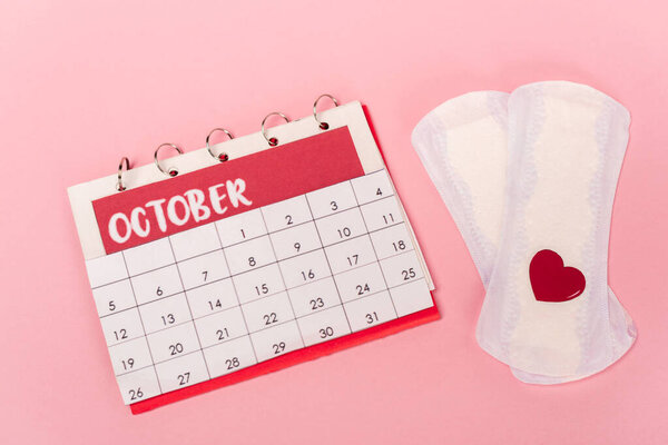 High angle view of feminine pads with paper heart and calendar on pink background