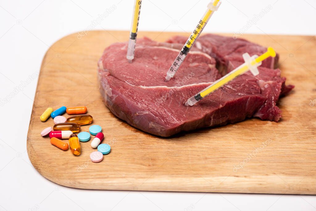 Syringes in raw meat and hormonal pills on cutting board isolated on white