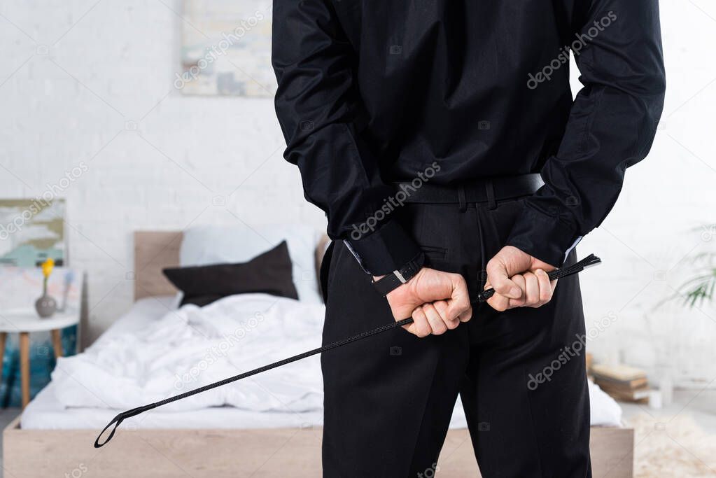 Cropped view of man with flogging whip near bed in bedroom