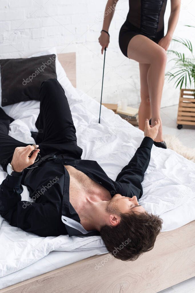 Selective focus of woman with flogging whip and man lying on bed with outstretched hand in bedroom