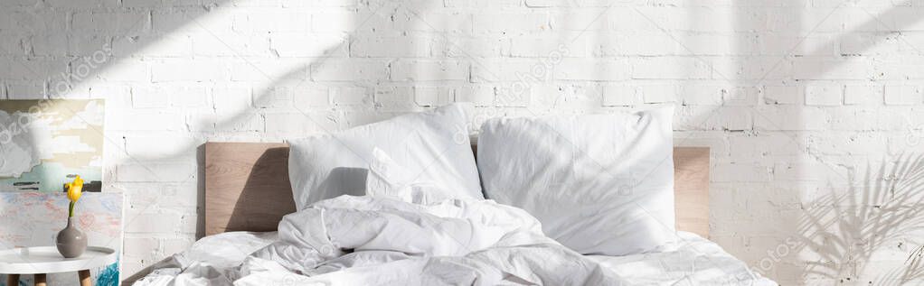 Bed near coffee table with vase and painted pictures near brick wall in bedroom, panoramic shot