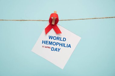 Card with world hemophilia day lettering and red ribbon on cord with pin isolated on blue  clipart