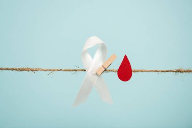 White ribbon with pin and paper in blood drop shape on rope isolated on blue, hemophilia concept clipart