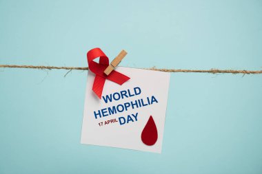 Card with world hemophilia day lettering and red ribbon on rope with pin isolated on blue  clipart