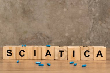 Selective focus of sciatica lettering on cubes with pills on wooden table on grey background clipart