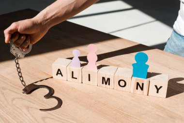 cropped view of man holding handcuffs near wooden blocks with alimony lettering and paper people on desk clipart