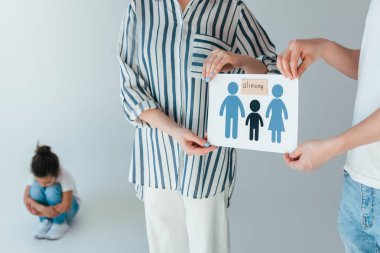selective focus of parents holding paper with drawn family and alimony lettering near upset african american daughter on white clipart