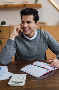 İş adamı gülümsüyor, başka tarafa bakıyor ve ofisteki masada akıllı telefondan konuşuyor.