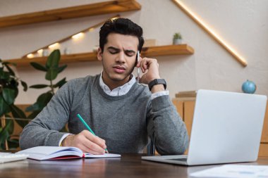 Akıllı telefondan konuşan ve masadaki dizüstü bilgisayarın yanında yazan bir iş adamı.