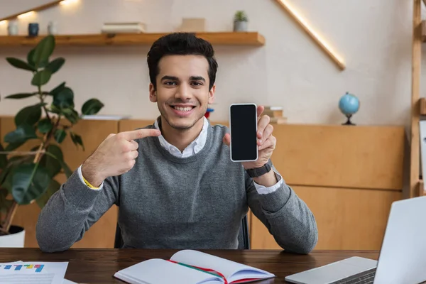 İş adamı kameraya bakıyor, gülümsüyor ve ofisteki masada akıllı telefonu işaret ediyor.