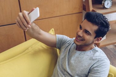 Adam gülümsüyor ve kanepede akıllı telefonuyla selfie çekiyor.