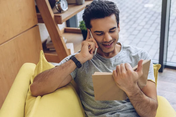 Vista Alto Ângulo Homem Com Livro Sorrindo Falando Smartphone Sofá — Fotografia de Stock