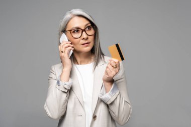 Güzel Asyalı iş kadını, gri saçlı, akıllı telefonuyla konuşan ve kredi kartını gri üzerinde tutan.
