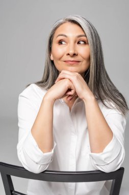 positive pensive asian woman with grey hair sitting on chair isolated on grey clipart