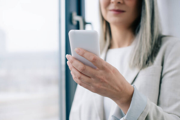 cropped view of businesswoman with grey hair using smartphone in office 