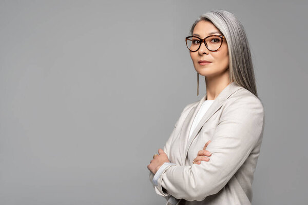 attractive asian businesswoman in eyeglasses with crossed arms isolated on grey
