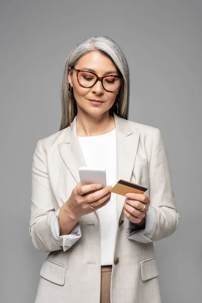 attractive asian businesswoman with grey hair shopping online with credit card and smartphone isolated on grey