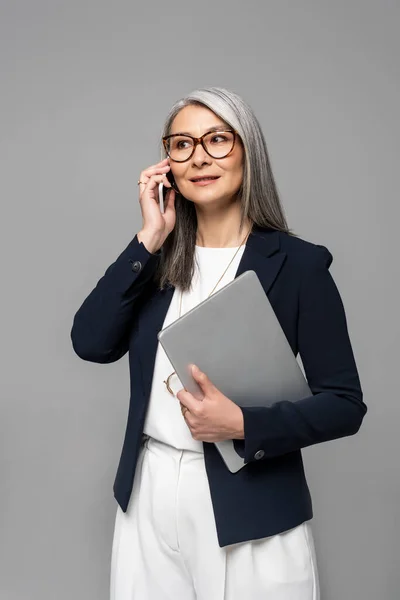 有魅力的亚洲女商人 头发灰白 在智能手机上说话 手里拿着用灰色隔离开的笔记本电脑 — 图库照片