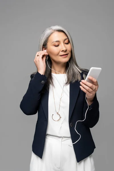 beautiful asian businesswoman with grey hair listening music with earphones and smartphone isolated on grey