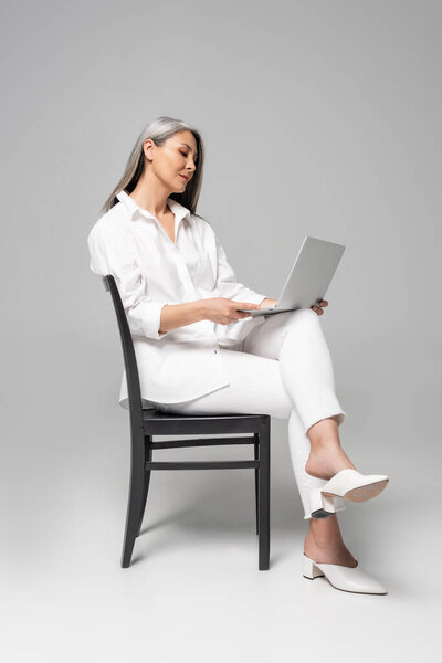 adult attractive asian woman with grey hair sitting on chair and using laptop on grey