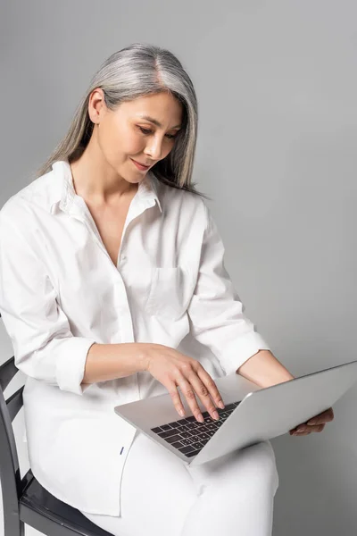 Улыбающаяся Азиатская Женщина Седыми Волосами Сидящая Стуле Использующая Ноутбук Сером — стоковое фото