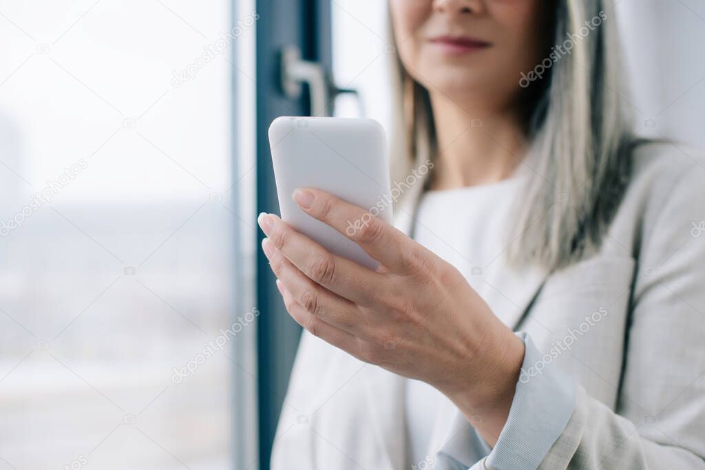 cropped view of businesswoman with grey hair using smartphone in office 