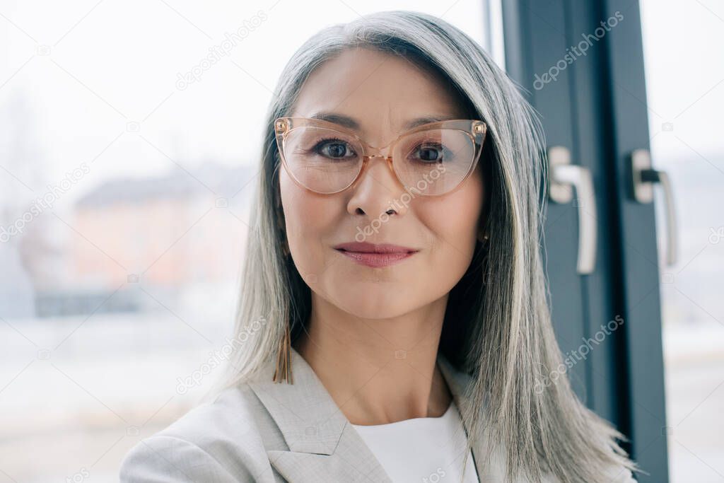 attractive confident asian businesswoman with grey hair in grey suit and eyeglasses looking at camera