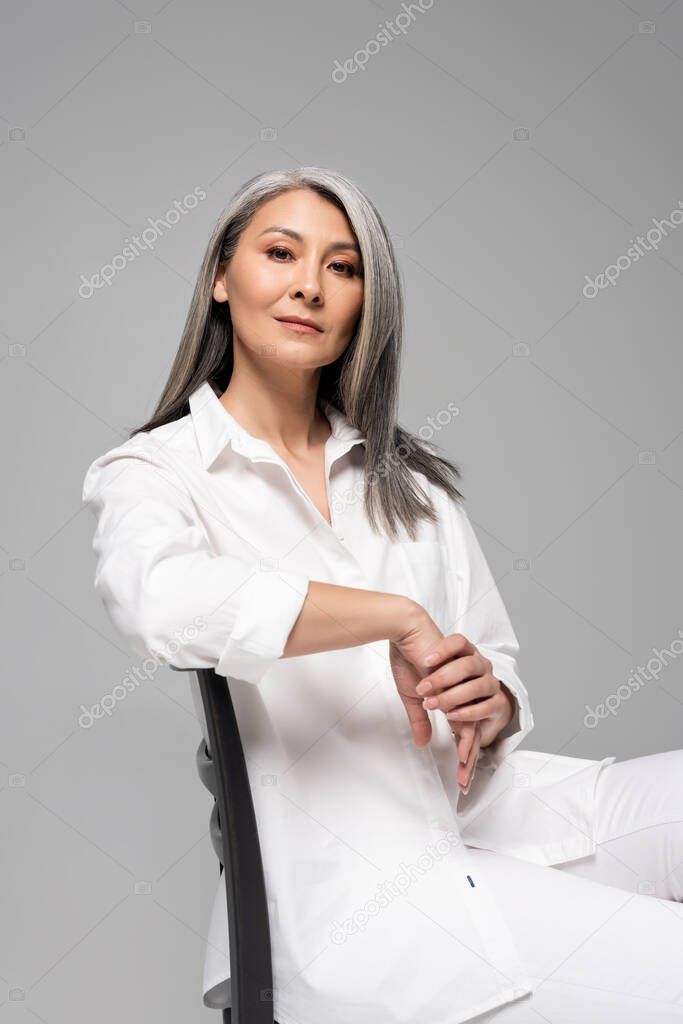 beautiful confident asian woman with grey hair sitting on chair isolated on grey