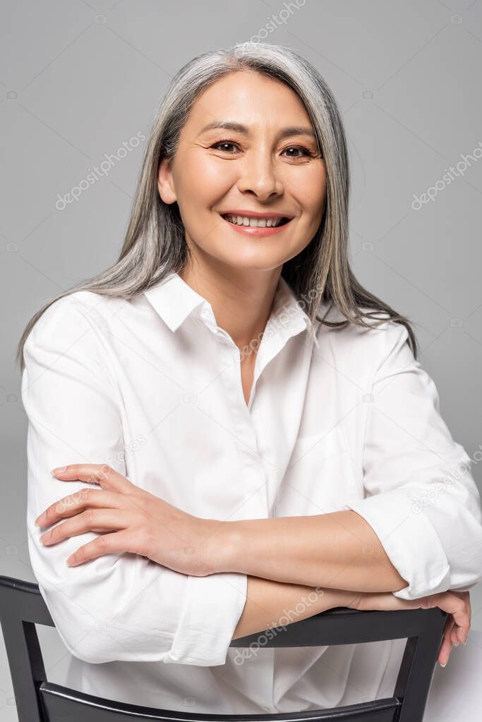 beautiful smiling asian woman with grey hair sitting on chair isolated on grey