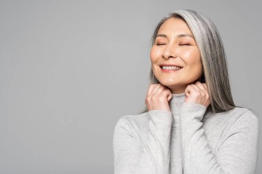happy asian woman in turtleneck with grey hair and closed eyes isolated on grey clipart