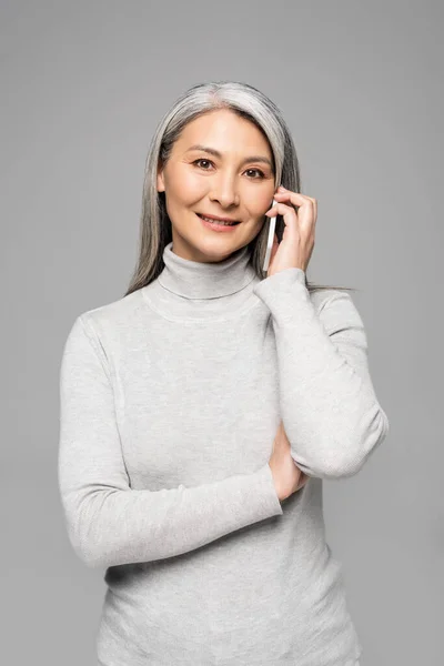 快乐的亚洲女人 穿着高领毛衣 灰白的头发 用智能手机与灰色隔离说话 — 图库照片