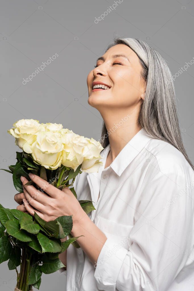 adult cheerful asian woman with grey hair holding bouquet of white roses isolated on grey