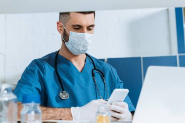 Laptop 'un yanında akıllı telefon kullanan tıbbi maskeli ve lateks eldivenli genç bir doktor.