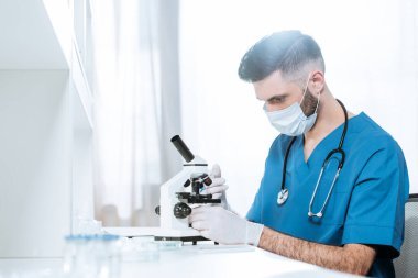 Laboratuvarda mikroskopla çalışan tıbbi maskeli ve lateks eldivenli genç bir biyolog.