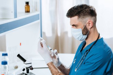 Tıbbi maskeli ve lateks eldivenli genç bir biyolog şırıngayla ilaç alıyor.