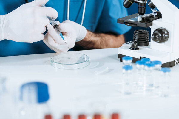 partial view of biologist in latex gloves holding syringe near petri dish